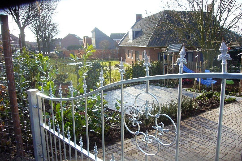 Een sierlijke metalen poort die toegang geeft tot een woontuin, met een huis en bomen op de achtergrond.