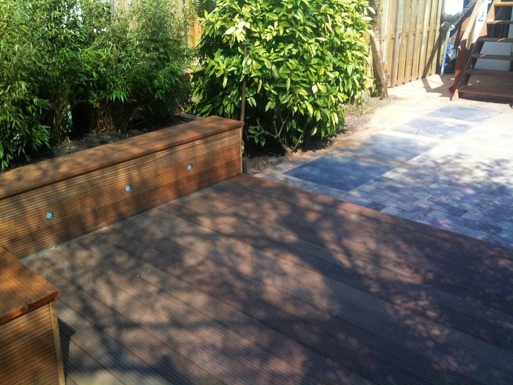 Houten terras met ingebouwde bank, grenzend aan een bloembed met groene struiken. Een verhard pad leidt ernaartoe.