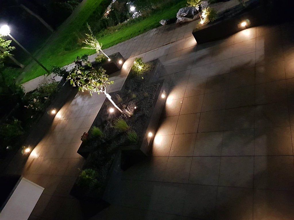 Een terras 's avonds, verlicht door schijnwerpers en een lantaarnpaal, met plantenbakken in verschillende niveaus en diverse beplanting.