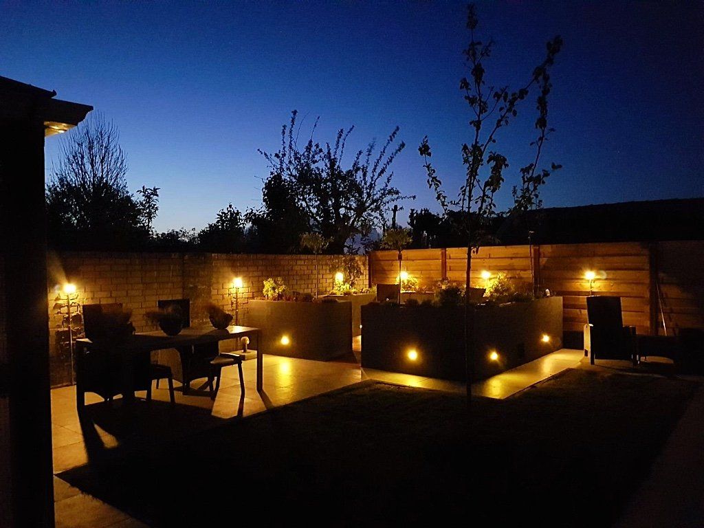 Terras in de achtertuin 's avonds met sfeerverlichting. Plantenbakken, zitgedeelte en houten schutting. Donkerblauwe lucht.