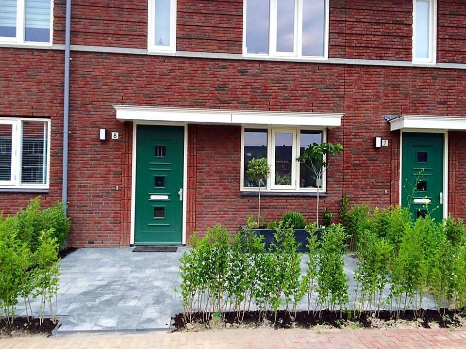 Rij bakstenen rijtjeshuizen met groene deuren en ramen, struiken ervoor.