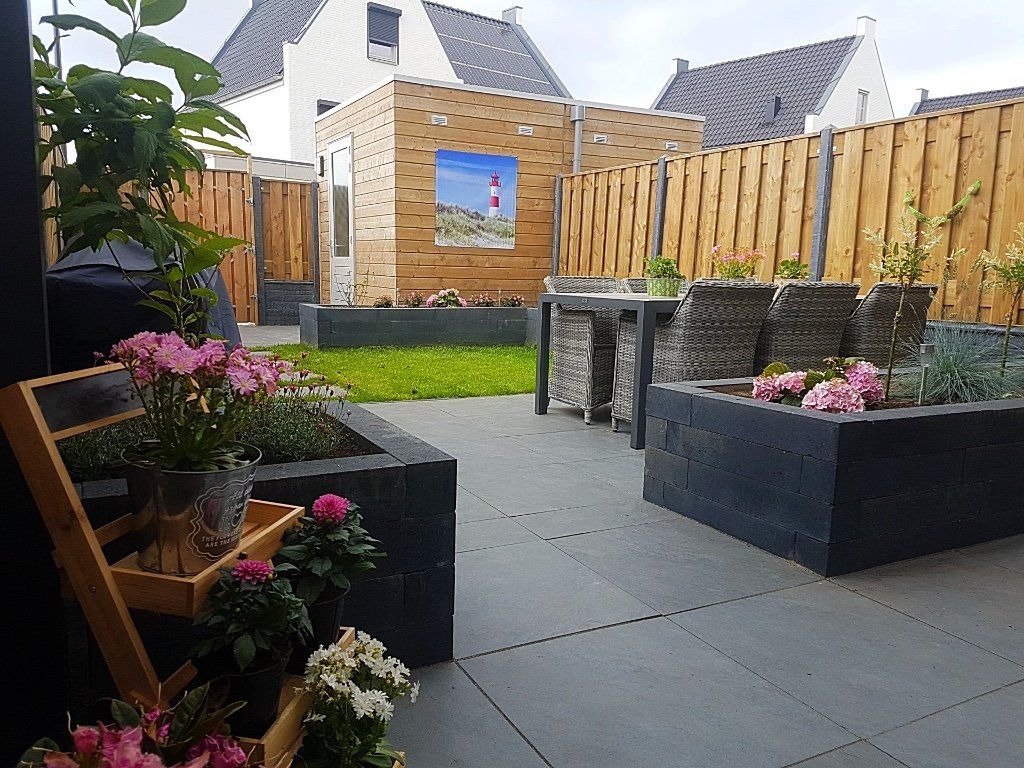 Terras in de achtertuin met grijze tegels, houten schutting, gras en tuinmeubilair. Bloembakken met bloemen.