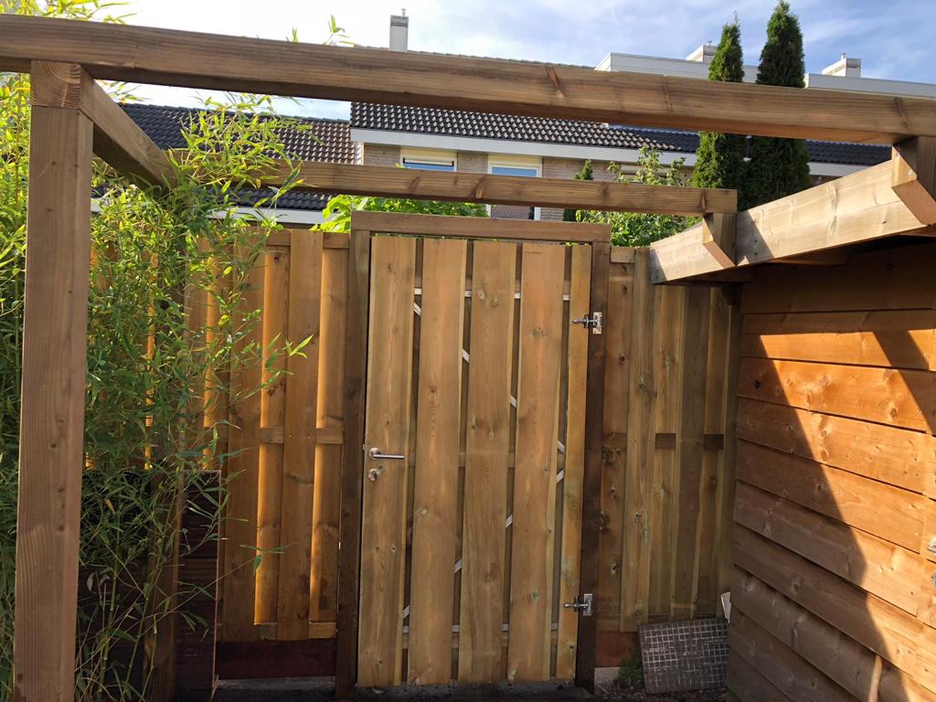 Houten poort en hek in een tuin, omgeven door groen en met houten balken aan het plafond.