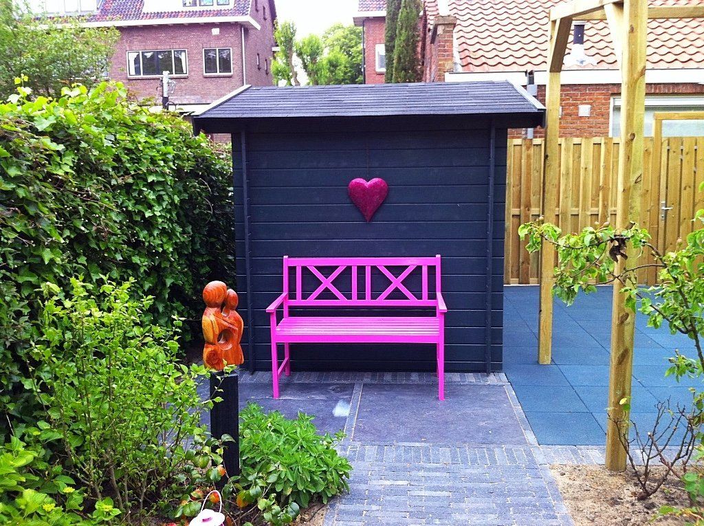 Een roze bankje staat voor een donker schuurtje met een hartvormige versiering in een achtertuin.