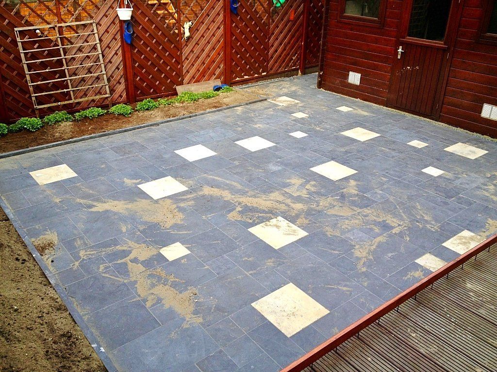 Terras bestraat met blauw-witte tegels, met vuilstrepen over het oppervlak, naast een houten schuur en een schutting.