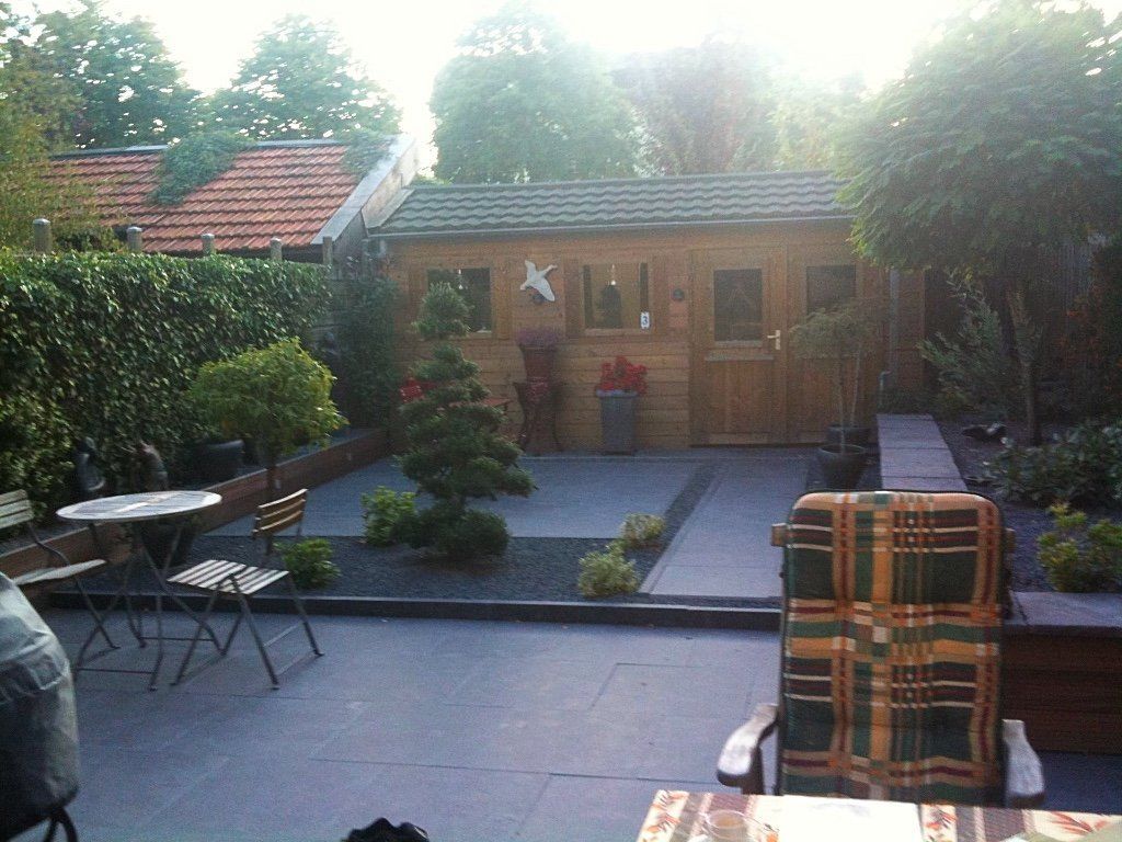 Houten schuur in een tuin met terras, kleine bomen en een stoel.