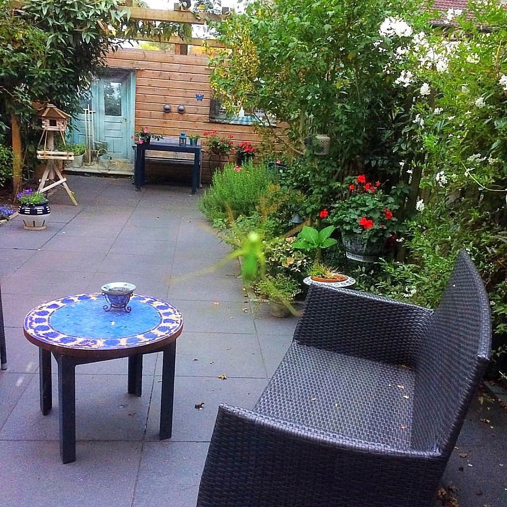 Een tuinterras met een mozaïektafel, rieten zitmeubelen en een bakstenen gebouw op de achtergrond.