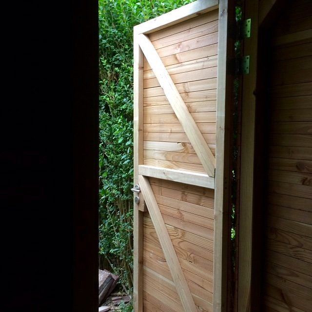 Een houten poort, gedeeltelijk open, staat in een tuin met groen gebladerte.