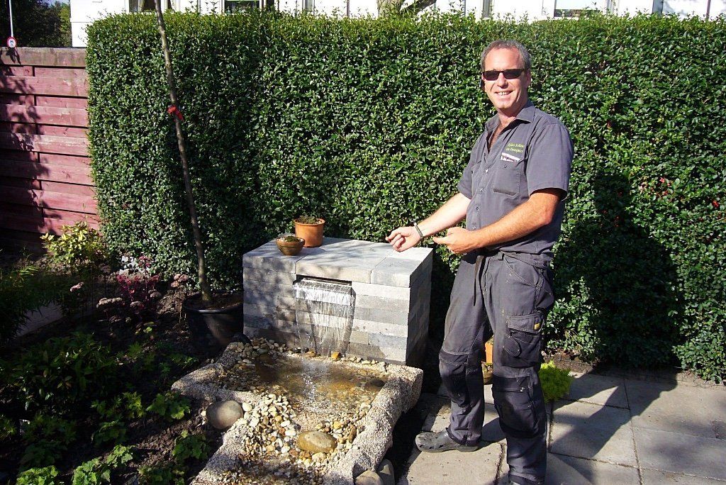 Een man wijst glimlachend naar een kleine stenen waterval in een tuin. Op de achtergrond is een groene heg te zien.