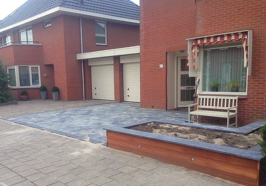 Bakstenen huis met garage en oprit; bankje onder het raam met zonnescherm; bloembed.