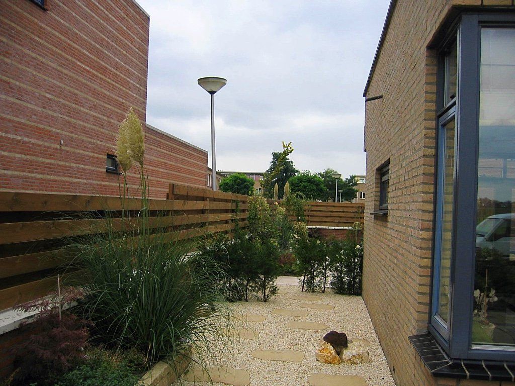 Smalle buitenruimte tussen bakstenen gebouwen, grindpad, houten schutting, groen, bewolkte lucht.