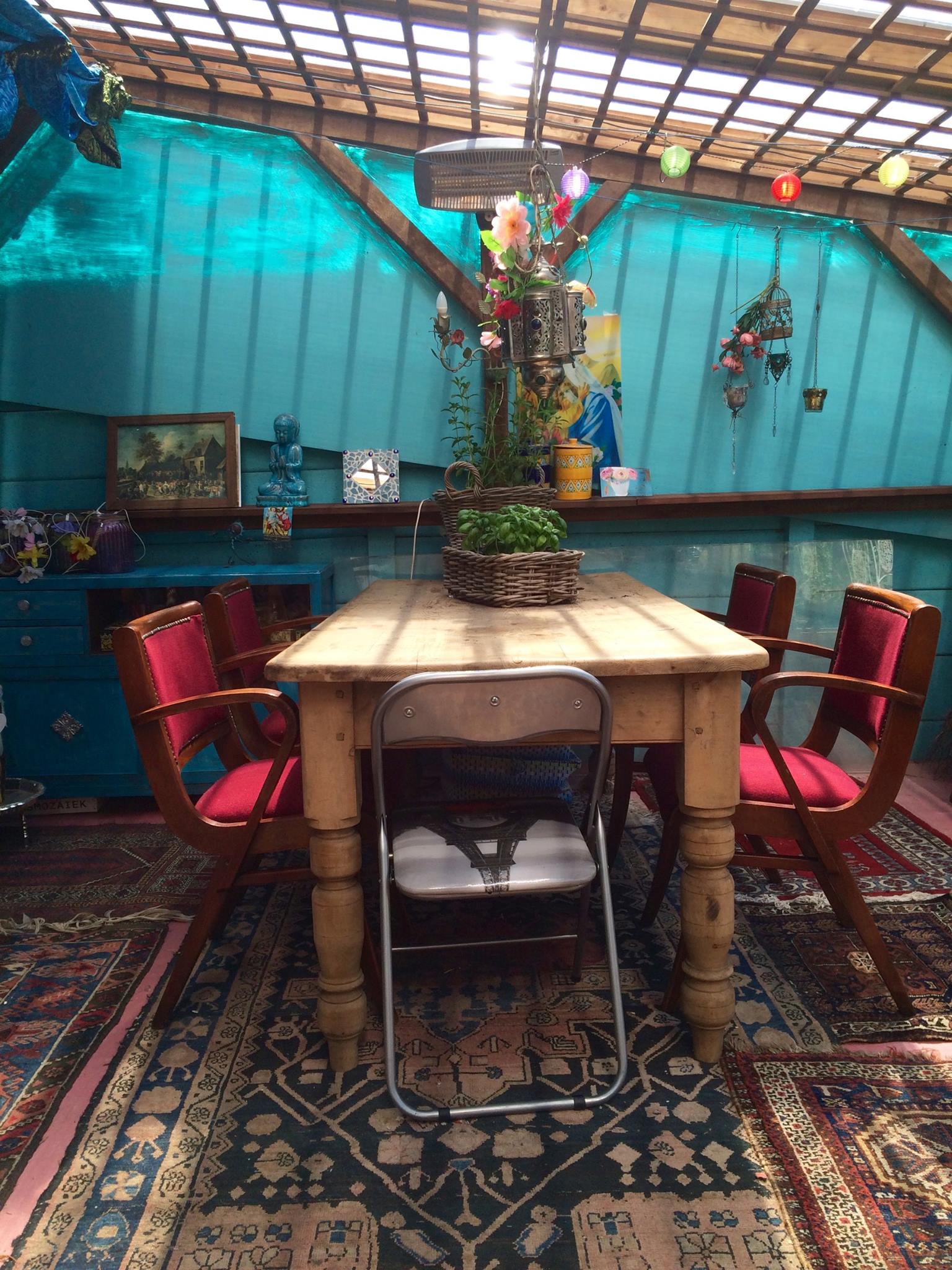 Eethoek: Houten tafel met rode stoelen, een klapstoel. Turquoise muur, vloerkleden en een plantenarrangement.