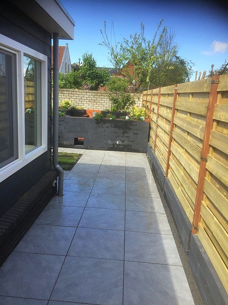 Terras met grijze tegels, naast een huis en een houten schutting, op een zonnige dag.