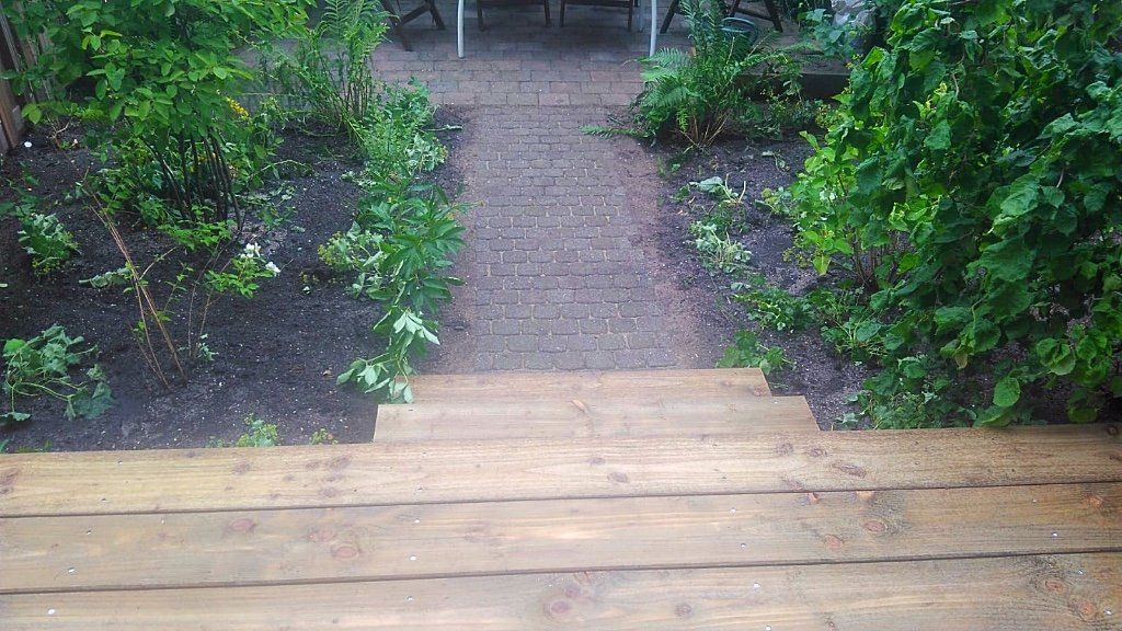 Houten treden leiden naar een tuinpad, geflankeerd door groene struiken, met bovenaan zichtbaar tuinmeubilair.