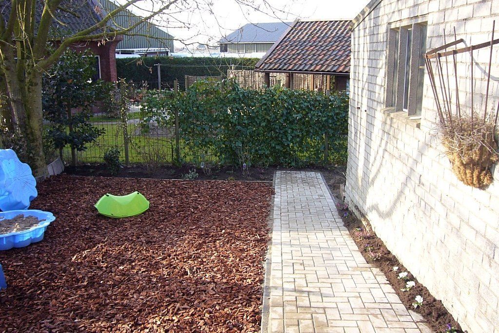 Achtertuin met bakstenen pad, mulch als bodembedekking, groene haag, wit gebouw en hangende bloemenmand.