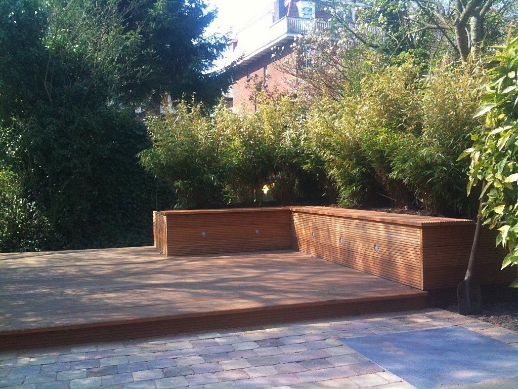 Houten terras met ingebouwde bank, omgeven door groen en met een gebouw op de achtergrond.
