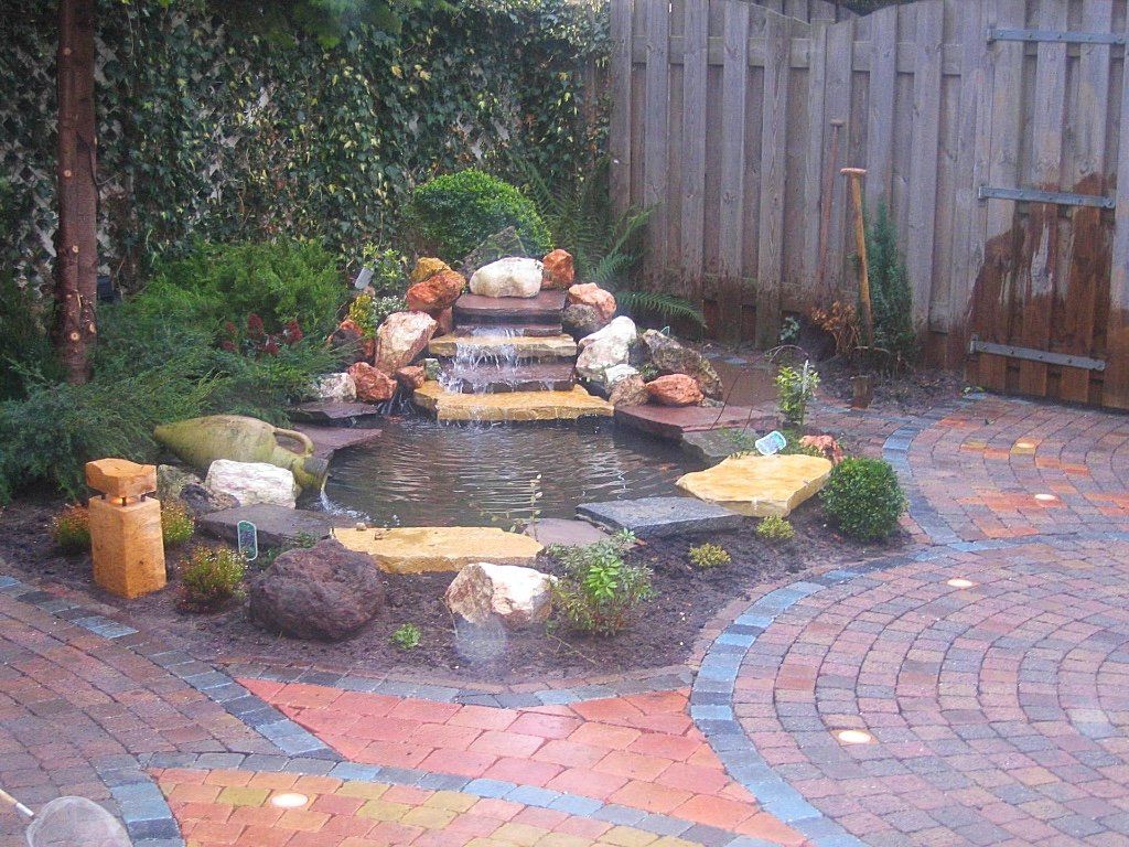 Kleine vijver in de achtertuin met een waterval, omgeven door rotsen, planten en bakstenen bestrating.