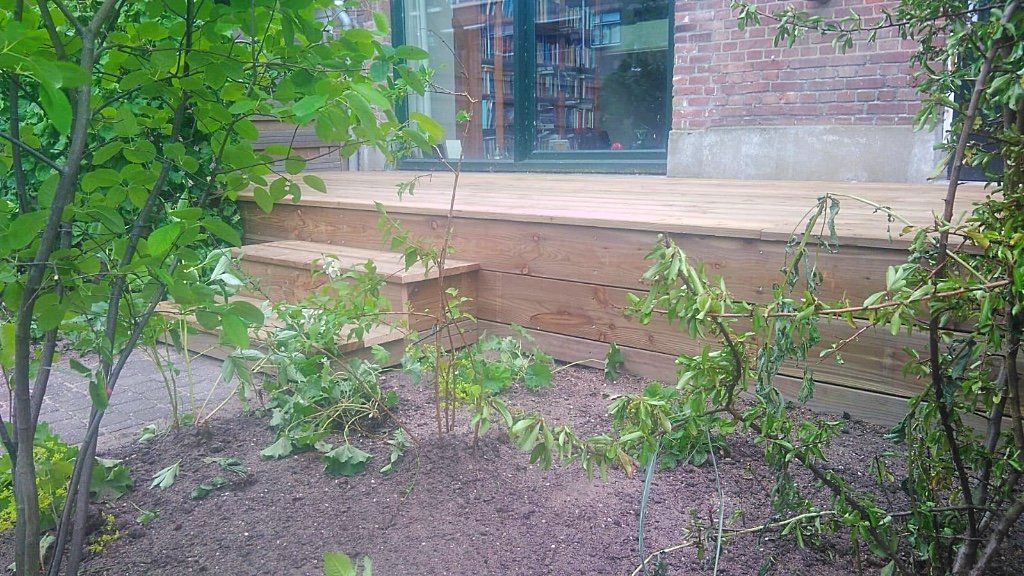 Houten terras met ingebouwde trap, grenzend aan een tuin met groene planten en een gebouw op de achtergrond.