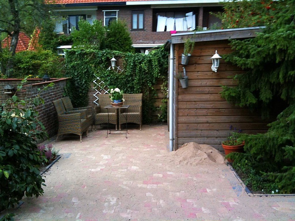 Terras in de achtertuin met bakstenen vloer, zitgedeelte, houten schuur en omringende beplanting.