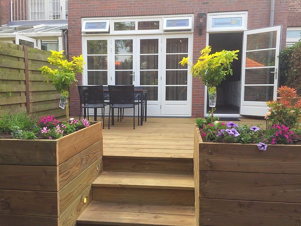 Houten terras en bloembakken in een achtertuin met een bakstenen huis en openslaande deuren.