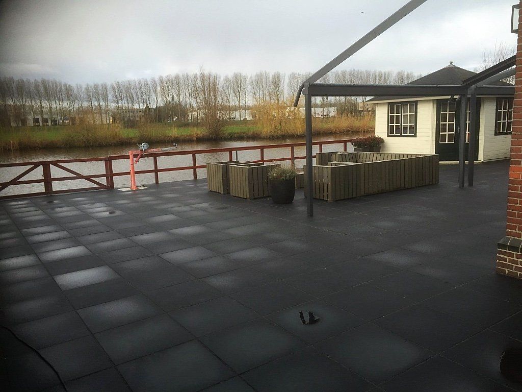 Buitenterras met donkere vierkante tegels, met uitzicht op een waterweg met een klein gebouw.