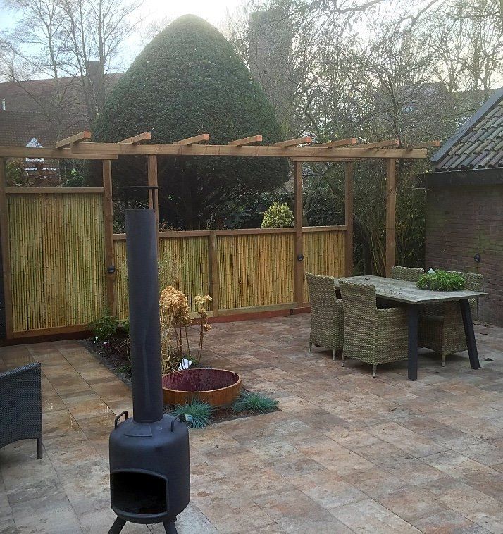 Terras met een pergola, een vlechtwerkscherm, een tafel met stoelen en een zwarte buitenhaard.