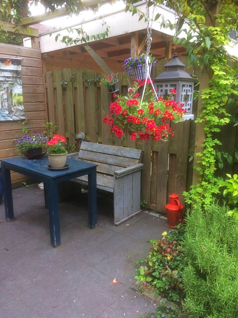 Gezellige zithoek buiten met tafel, bank en hangende rode bloemen bij een houten schutting.