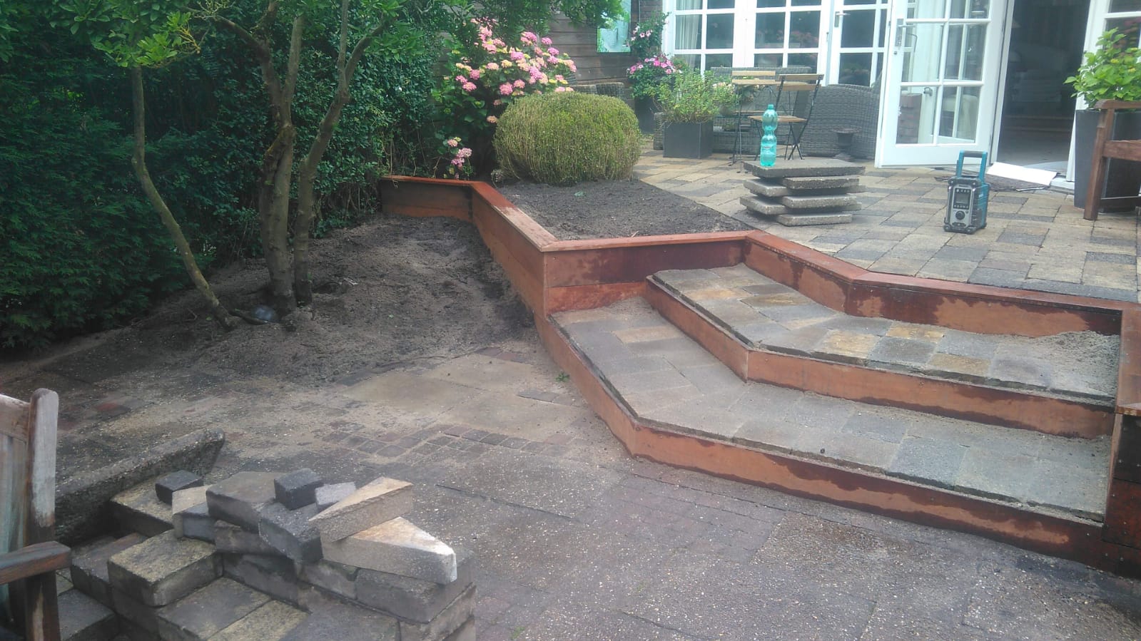 Een buitentrap van baksteen en hout leidt naar een terras met op de achtergrond een tuin en een huis.