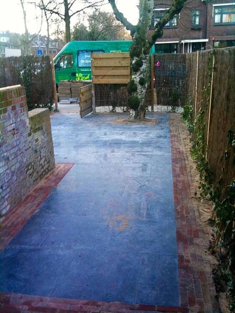 Een blauw geplaveid gebied, omgeven door bakstenen muren en houten hekken, met een boom in het midden. Op de achtergrond staat een groene bestelwagen.
