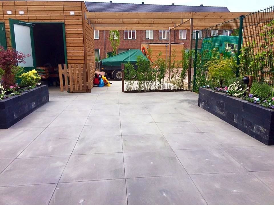 Buitenterras met grijze natuurstenen bestrating, houten constructies en bloembakken met planten.
