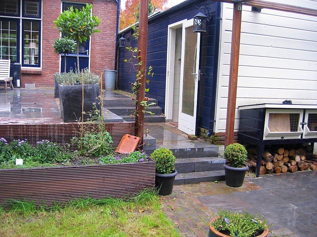 Achtertuin met een plantenbak van golfplaat, een trap naar een donkerblauwe constructie en een houtstapel.