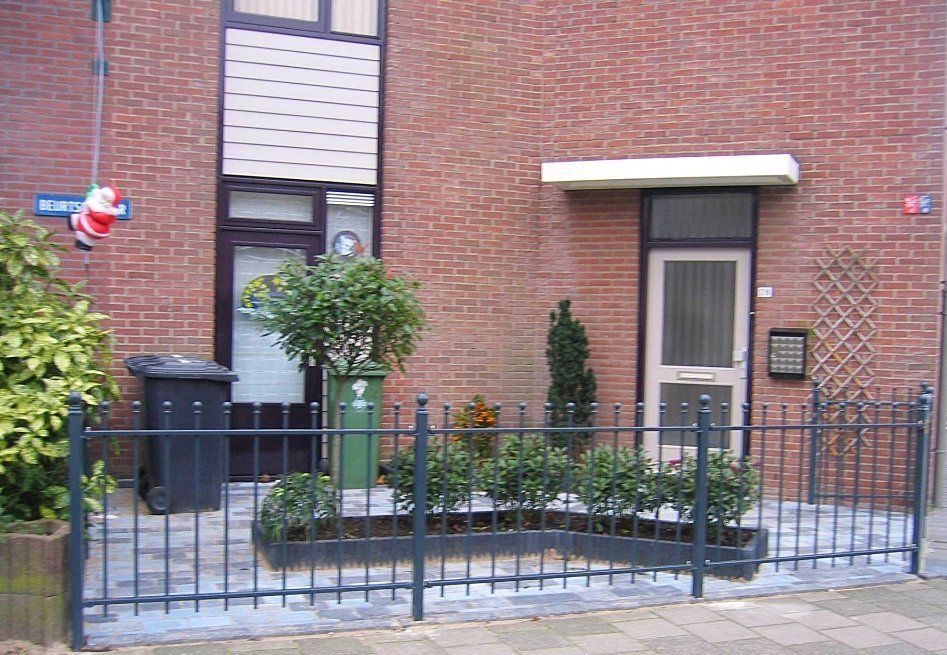 Een bakstenen gebouw met twee voordeuren, een hek en planten. Boven hangt een kerstmanversiering.