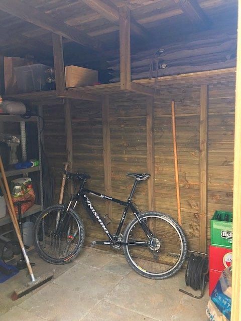 Een zwarte fiets staat in een houten schuur tegen de muur. Er zijn ook gereedschappen en opbergplanken aanwezig.