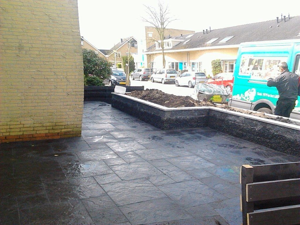 Zwart geplaveid terras met een lage, donkere keermuur en een beplante zone, straat op de achtergrond.