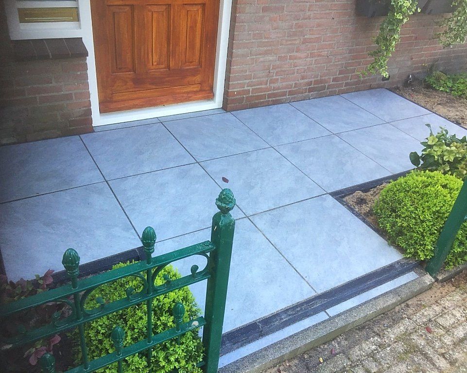 Grijze betegelde veranda met bruine deur, bakstenen muur en groen smeedijzeren hek.