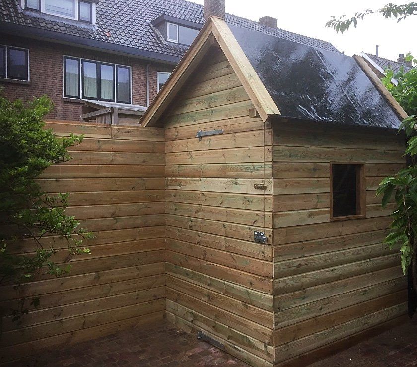 Houten schuur met een klein raam, gebouwd naast een houten schutting in een tuin.