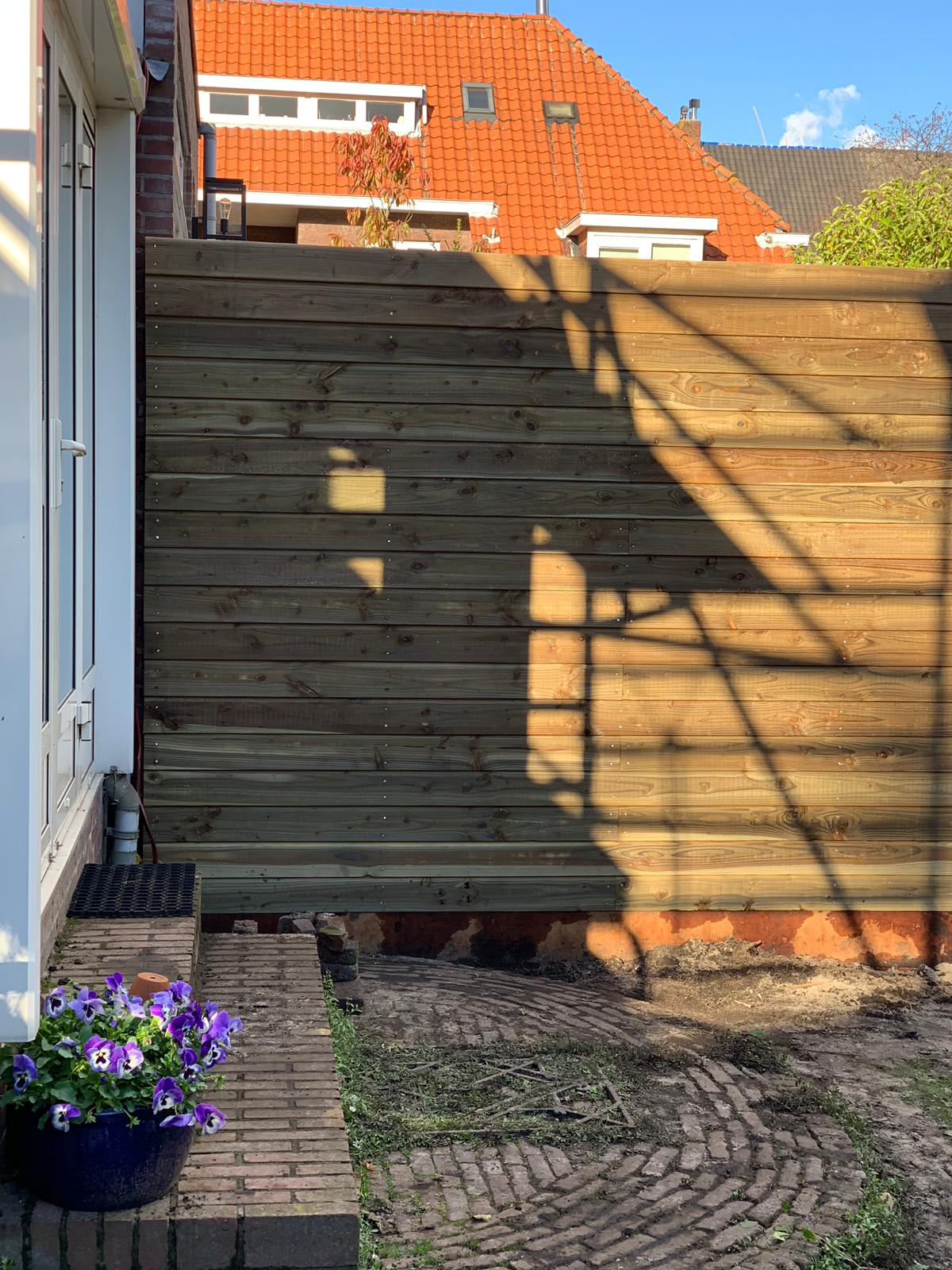 Houten schutting in de achtertuin, bakstenen muur, blauwe bloemen in een pot, rood pannendak op de achtergrond.