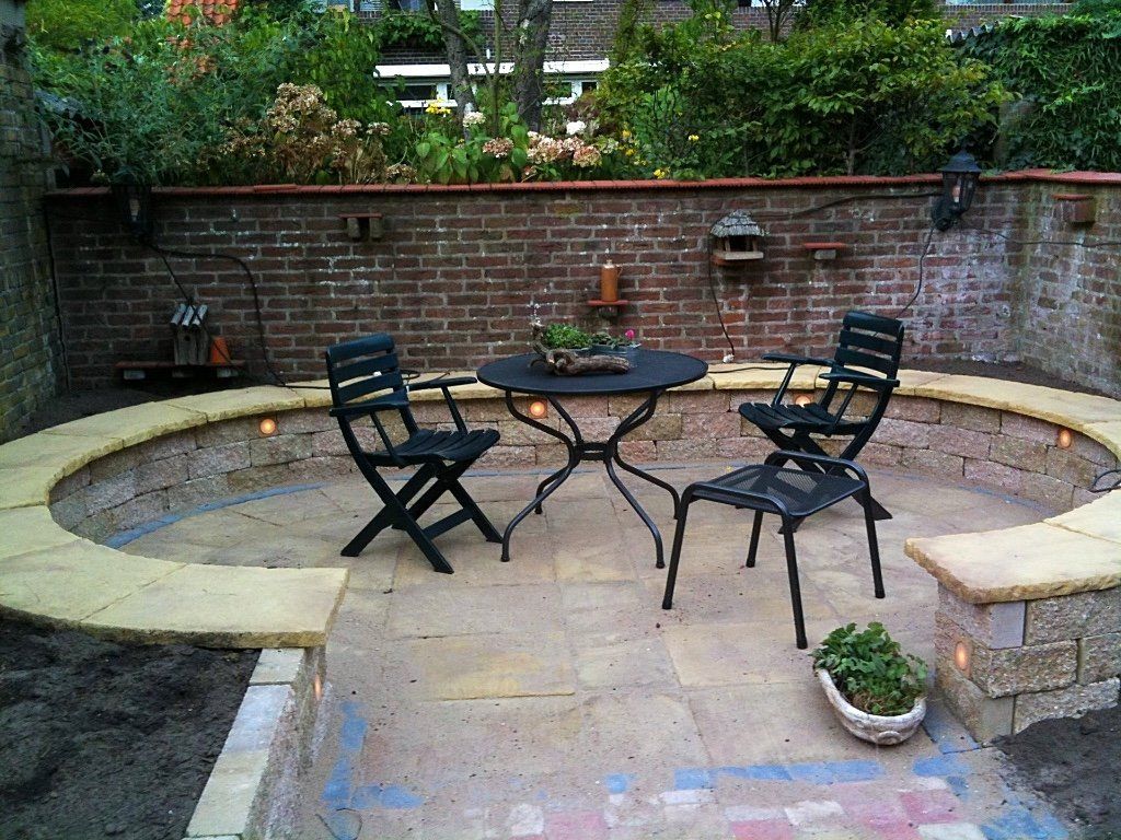 Terras met bakstenen muur, ronde stenen zitbank, zwarte metalen tafel en stoelen, en potplanten.