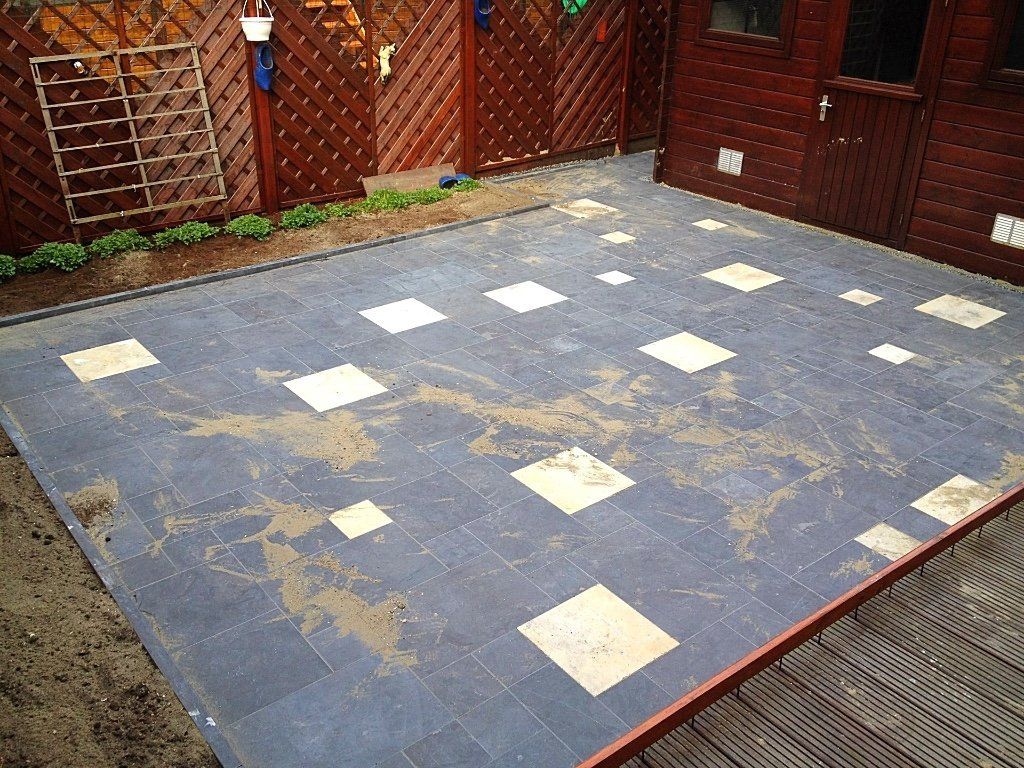 Terras met donkergrijze tegels, beige vierkanten, aarde en een houten schuur.