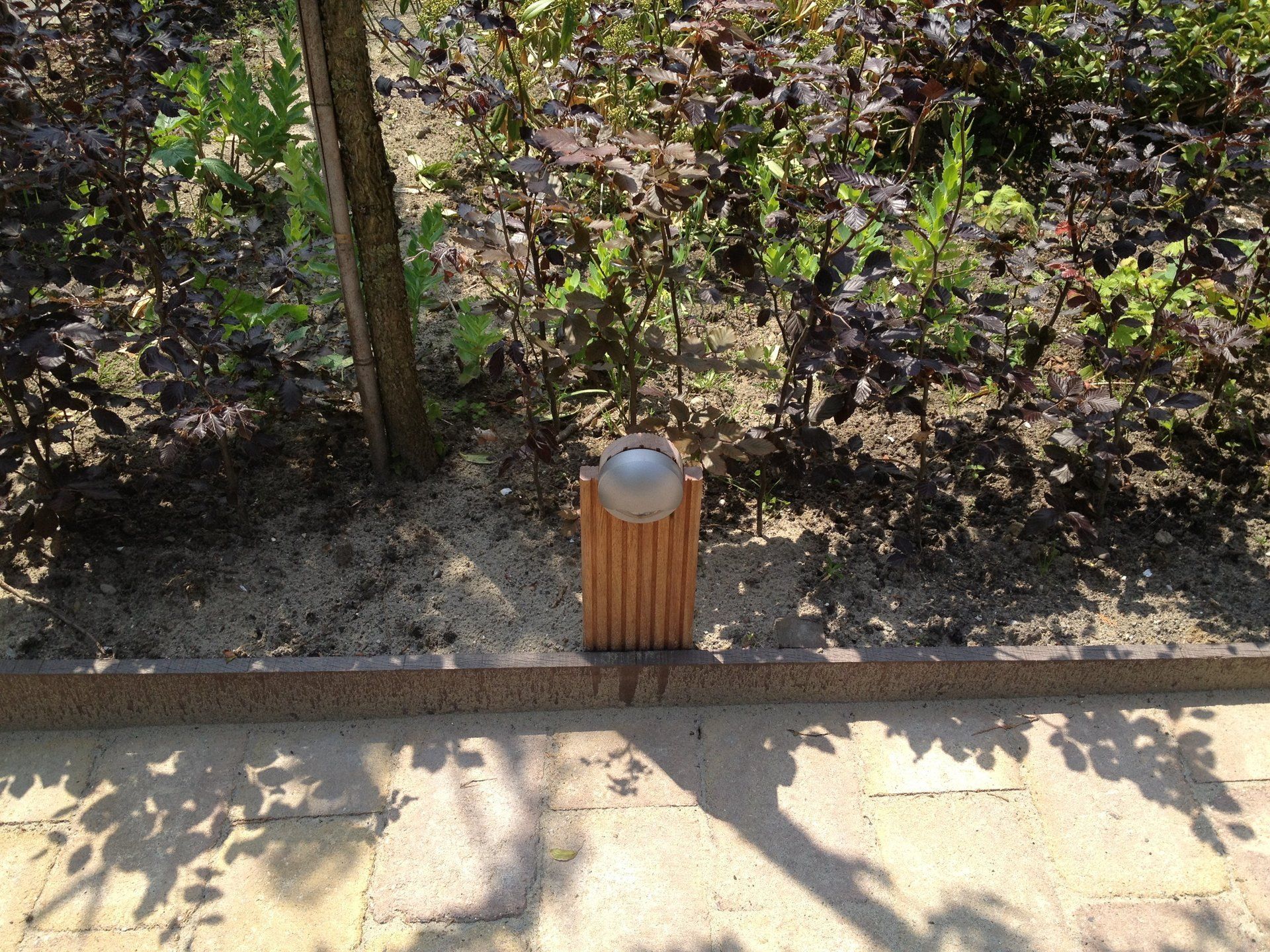 Een kleine metalen bol op een bruine houten steunconstructie in een bloembed met planten en een boom.