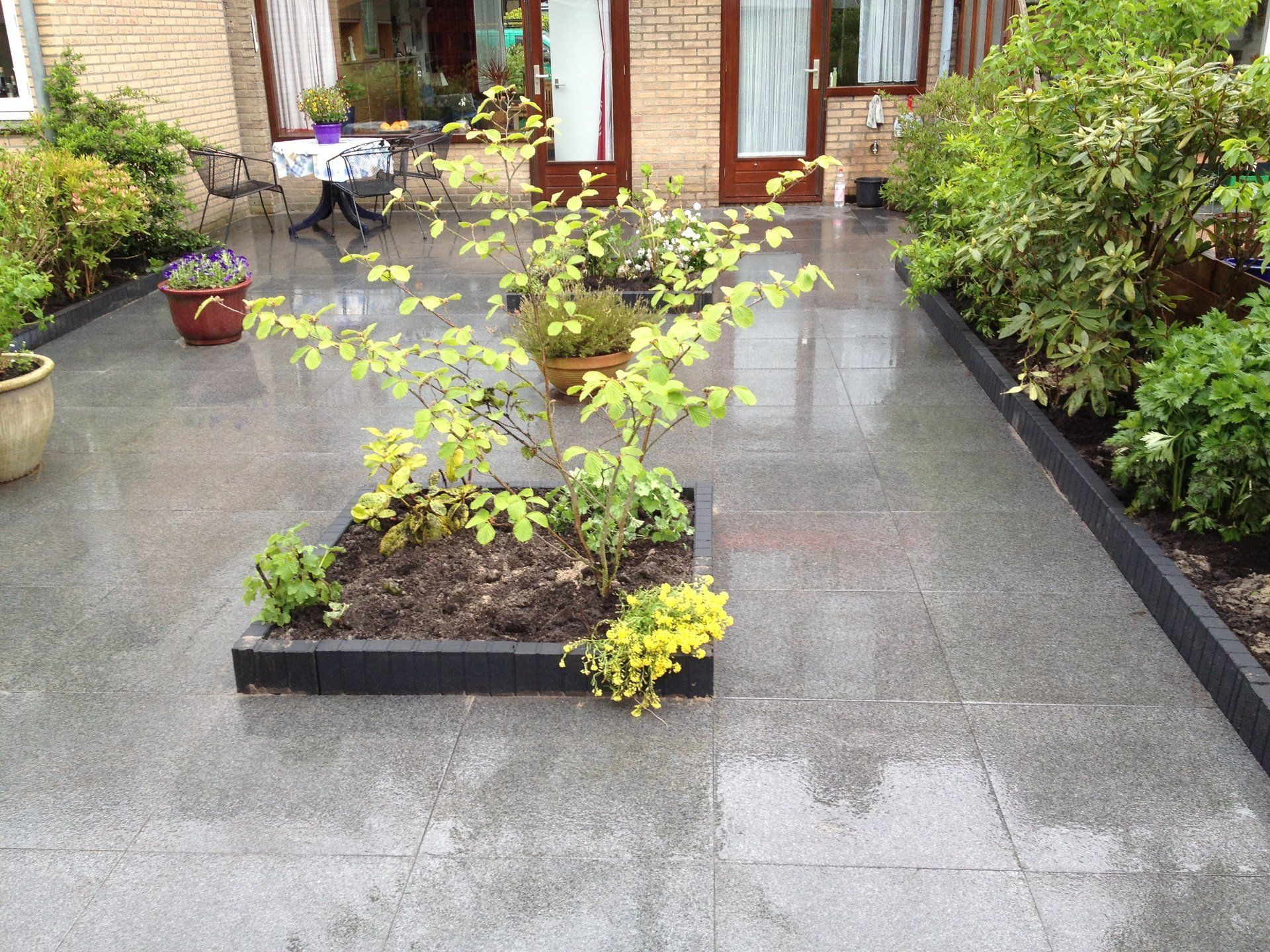 Een betegeld terras met een verhoogde plantenbak in het midden en omringende beplanting, nat van de regen.