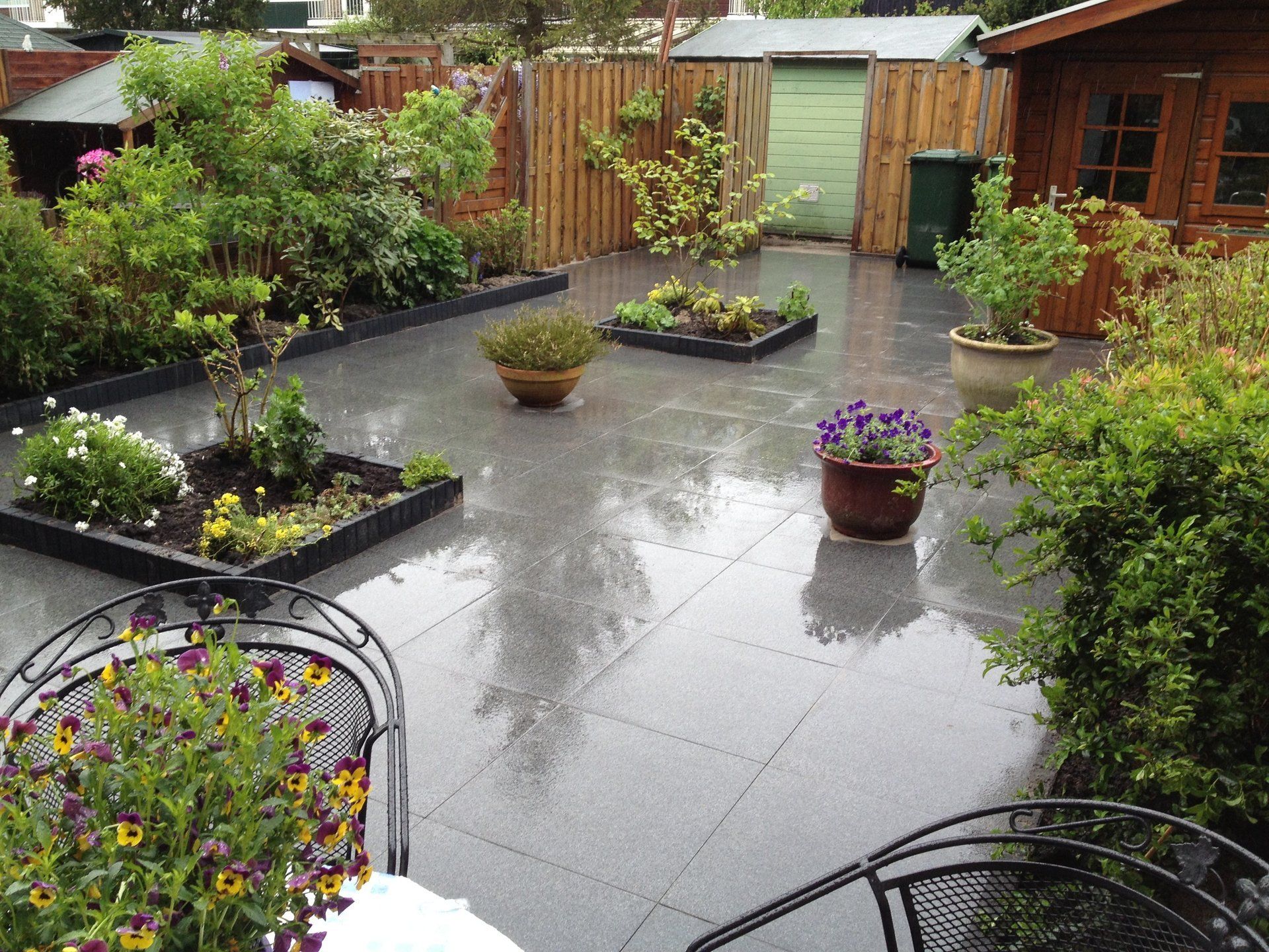 Een nat terras in de achtertuin met potplanten, bloemperken, een houten schutting en een schuur.