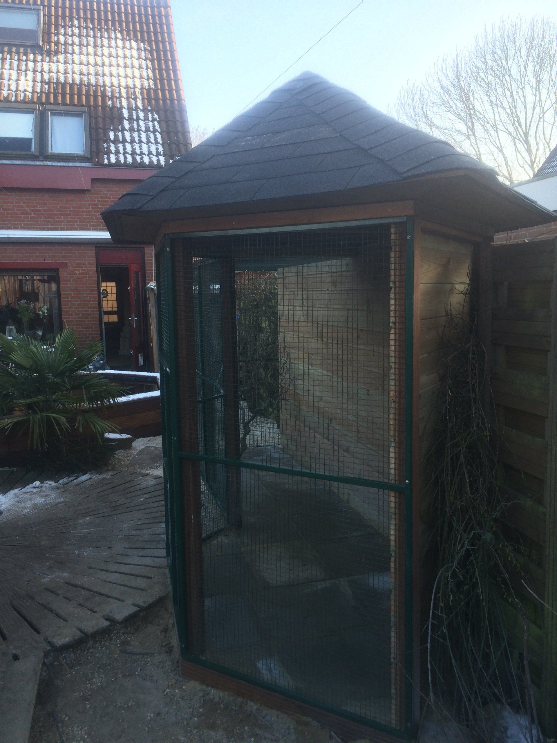 Buitenvolière met een donker dak en gaaswanden, op een stenen terras.