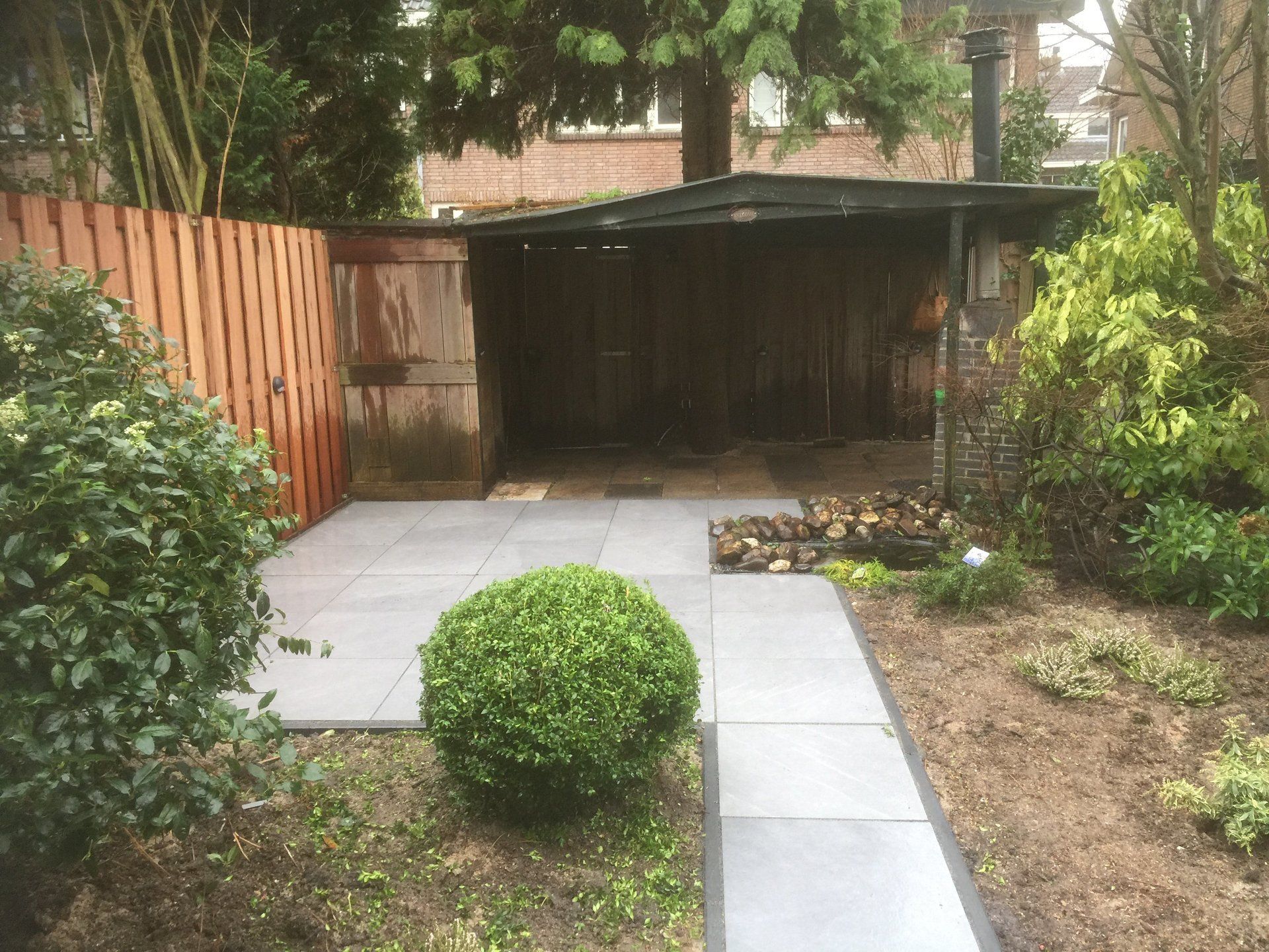 Achtertuin met grijs terras, kleine struik, pad, houten schuur en bruine schutting.