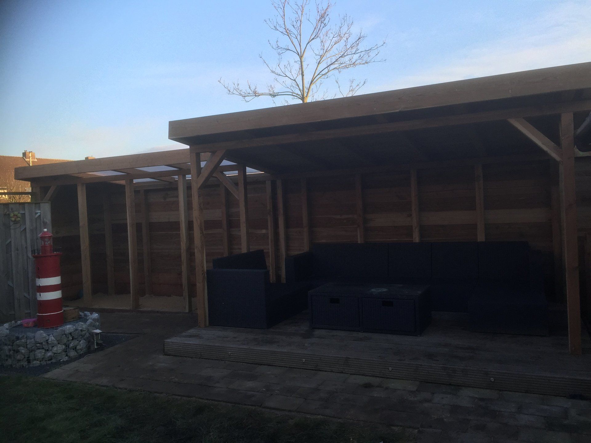 Houten buitenverblijf met zitgelegenheid, naast een stenen pad en een rood vuurtorenbeeld.