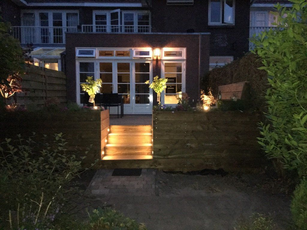 Achtertuin 's nachts met verlichte trappen die leiden naar een bakstenen gebouw met glazen deuren en ramen, en omgeven door groen.