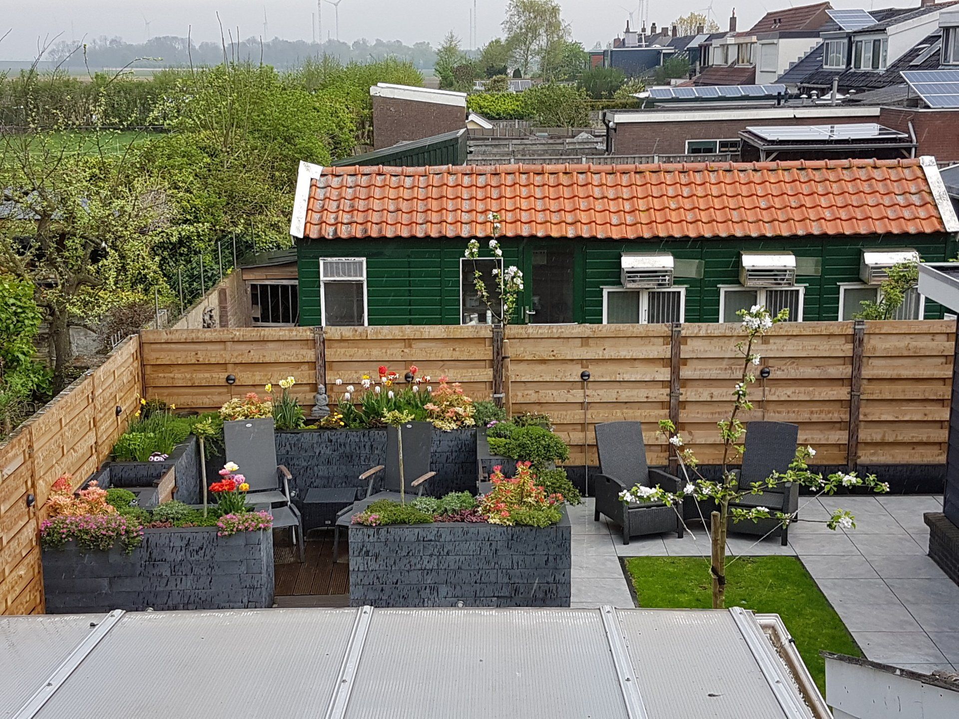 Achtertuin met houten schutting, plantenbakken, zitgelegenheid en een groene schuur met een oranje dak.