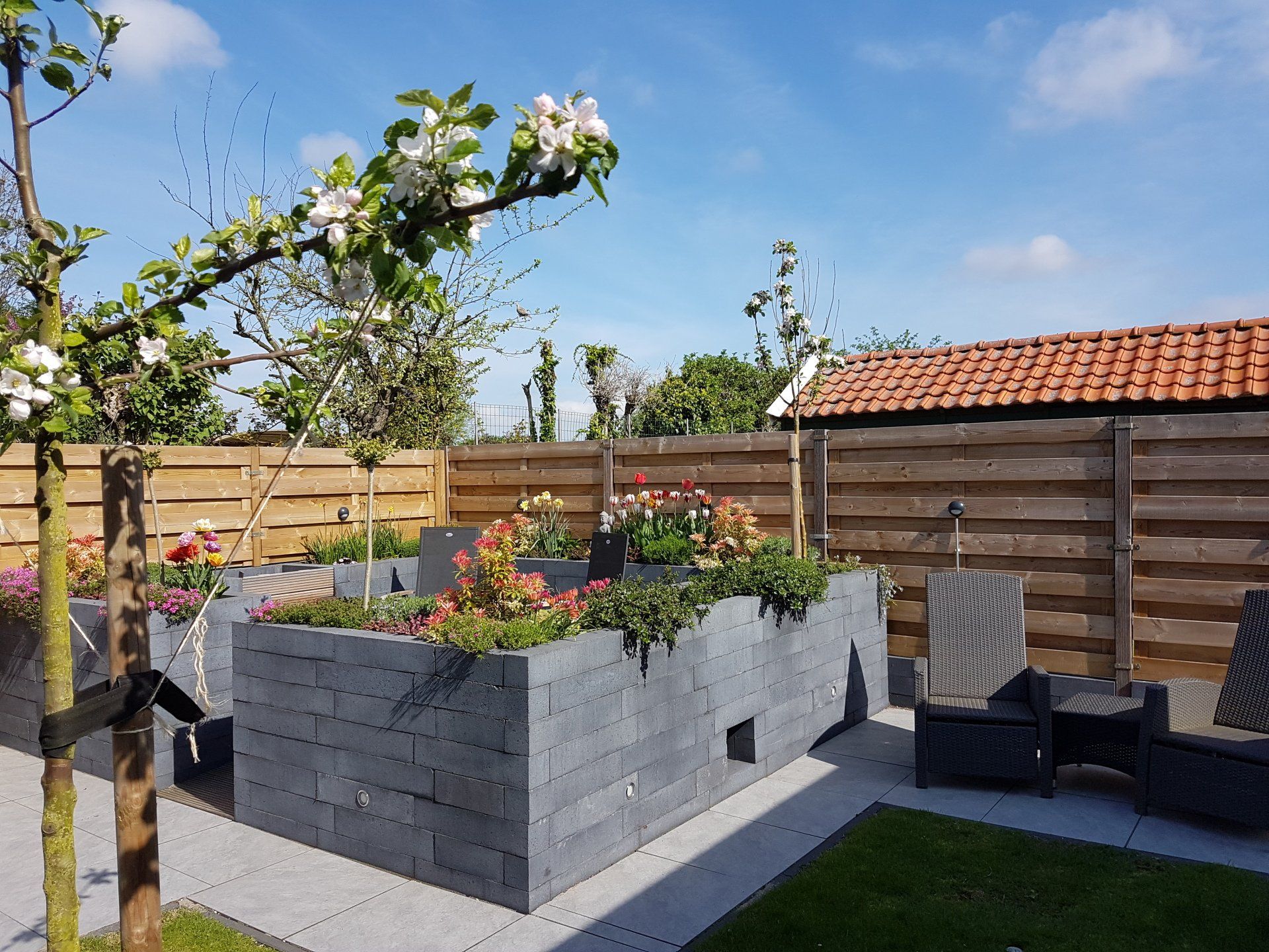Verhoogde bloemperken met kleurrijke bloemen, geflankeerd door een terras, gazon en houten schutting op een zonnige dag.