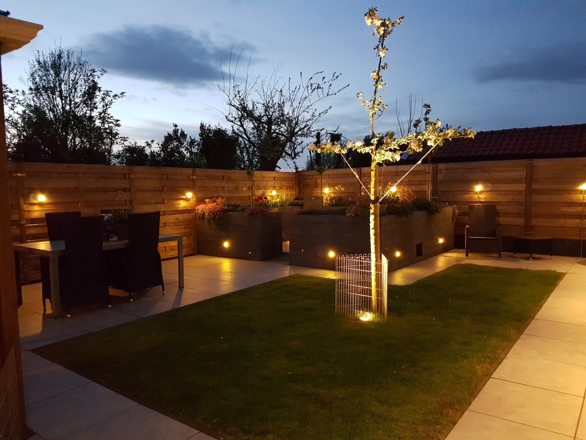 Terras in de schemering met verlichte plantenbakken, gazon en houten schutting.