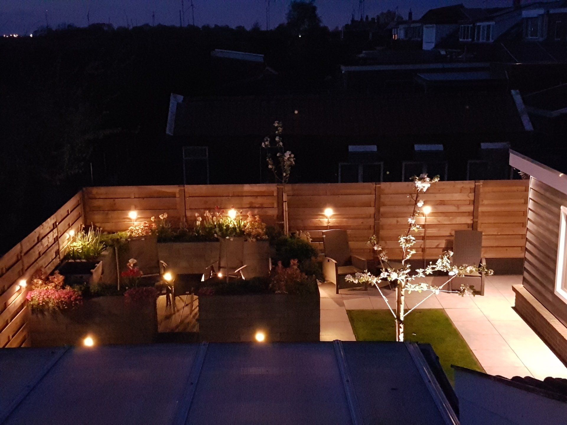 Een verlichte achtertuin 's nachts met een houten schutting, plantenbakken, een terras, zitjes en lichtslingers.